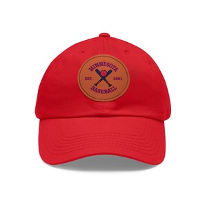 Op de afbeelding: Rode baseballpet met een bruine leren patch met een honkbal, gekruiste knuppels en de tekst "Minnesota Est. 1861 Baseball".