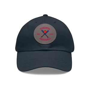 Op de afbeelding: Een donkerblauwe baseballpet met een ronde patch met een baseballlogo en de tekst "TORONTO EST. 1977 BASEBALL".