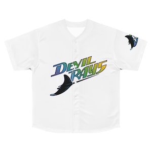 May include: White baseball jersey with a v-neck and button-up front. The jersey features the text "DEVIL RAYS" in a gradient of blue, green, and yellow, along with a black stingray graphic. A smaller stingray logo is on the sleeve.