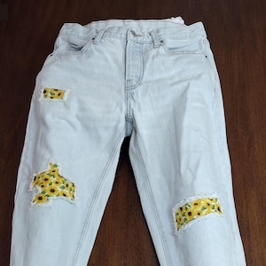 May include: Light-wash denim jeans featuring a button fly, belt loops, and five pockets. The jeans have three distressed areas with frayed edges, each backed with a yellow fabric patch featuring a repeating sunflower print pattern.
