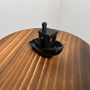 Op de afbeelding: Een klein, zwart, 3D-geprint model van een sleepboot met een schoorsteen op het dak. De boot staat op een houten oppervlak.
