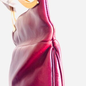 May include: A burgundy leather handbag with a gold-toned handle. The bag has a curved shape and a pleated design on the side. The handle is attached to the bag with gold-toned hardware.