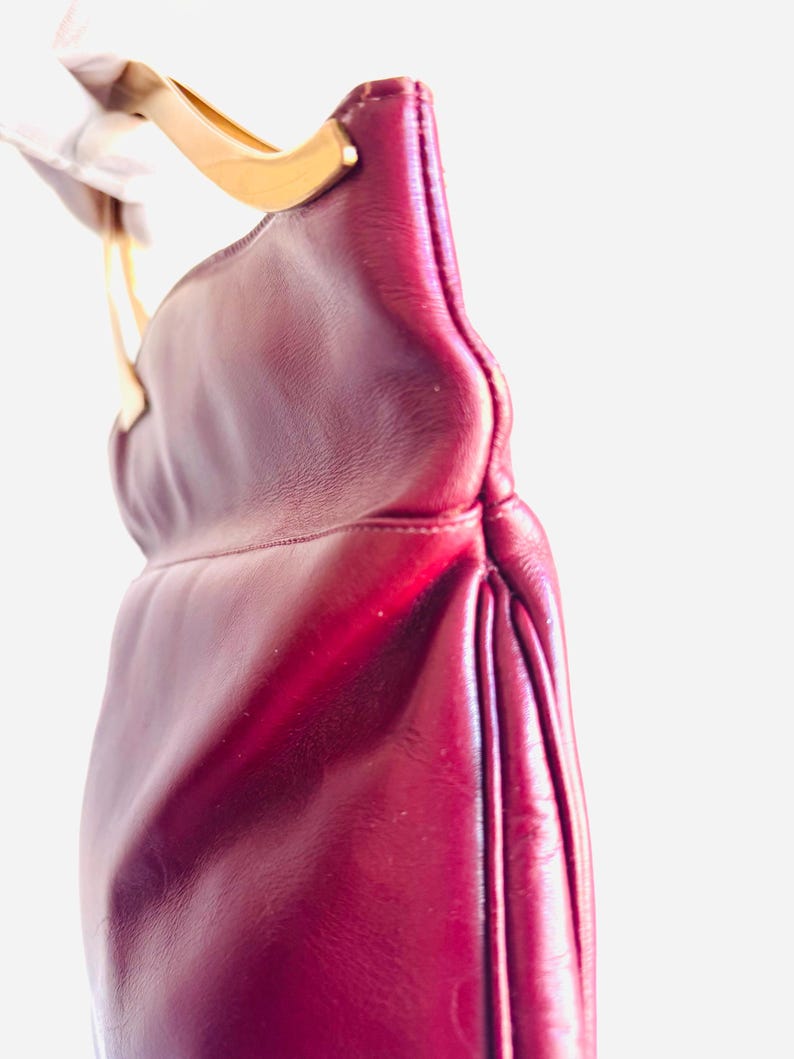 May include: A burgundy leather handbag with a gold-toned handle. The bag has a curved shape and a pleated design on the side. The handle is attached to the bag with gold-toned hardware.