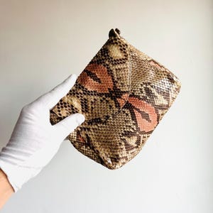 K&ouml;nnte beinhalten: Schlangenleder-Clutch in Braun-, Beige- und Korallt&ouml;nen. Die Clutch hat einen Klappverschluss und eine strukturierte Oberfl&auml;che. Sie wird von einer behandschuhten Hand vor einem schlichten Hintergrund gehalten.