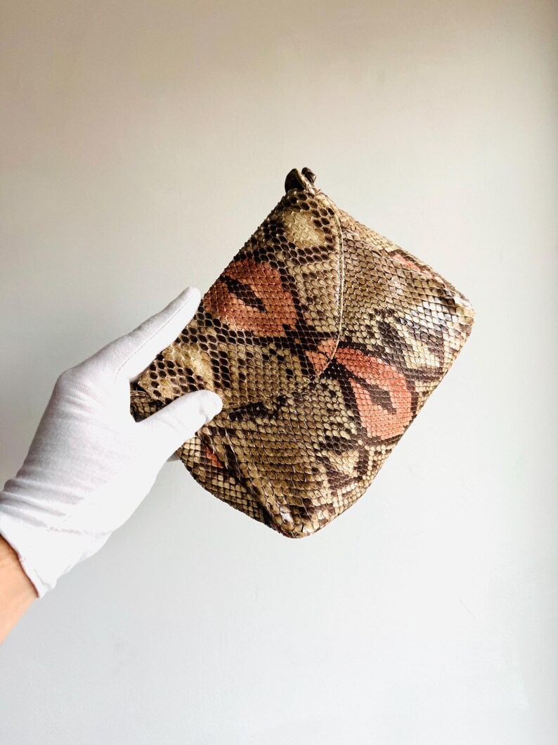K&ouml;nnte beinhalten: Schlangenleder-Clutch in Braun-, Beige- und Korallt&ouml;nen. Die Clutch hat einen Klappverschluss und eine strukturierte Oberfl&auml;che. Sie wird von einer behandschuhten Hand vor einem schlichten Hintergrund gehalten.