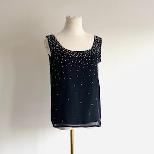 May include: A sleeveless black top featuring a scoop neckline and a sheer overlay. The garment is embellished with scattered silver-toned rhinestones that are densely concentrated around the neckline and become more sparse toward the hem.