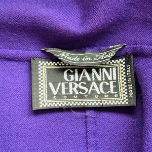 Pode incluir: Close-up de um tecido roxo com uma etiqueta preta. A etiqueta diz "GIANNI VERSACE COUTURE" em letras brancas, com o texto "Made in Italy". O tecido tem uma apar&ecirc;ncia texturizada.