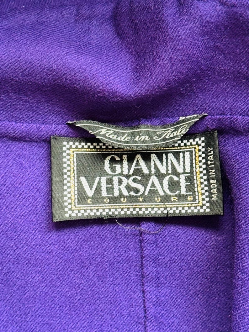Pode incluir: Close-up de um tecido roxo com uma etiqueta preta. A etiqueta diz "GIANNI VERSACE COUTURE" em letras brancas, com o texto "Made in Italy". O tecido tem uma apar&ecirc;ncia texturizada.