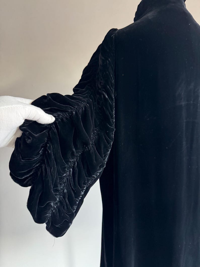 K&ouml;nnte beinhalten: Ein schwarzes Samtkleid mit langen &Auml;rmeln. Die &Auml;rmel sind gerafft und haben einen ger&uuml;schten Look.
