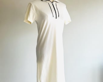 May include: Cream-colored short-sleeved dress with a collared neckline and a black trim. The dress features a zipper detail down the front and a knee-length hemline. The dress is displayed on a mannequin.