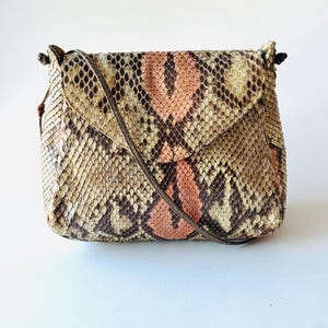 K&ouml;nnte beinhalten: Eine kleine, rechteckige Umh&auml;ngetasche mit Klappenverschluss. Die Tasche ist aus Schlangenhaut in den Farbt&ouml;nen Beige, Braun und Pfirsich gefertigt. Sie hat einen d&uuml;nnen, braunen Riemen und ein schlichtes, elegantes Design.