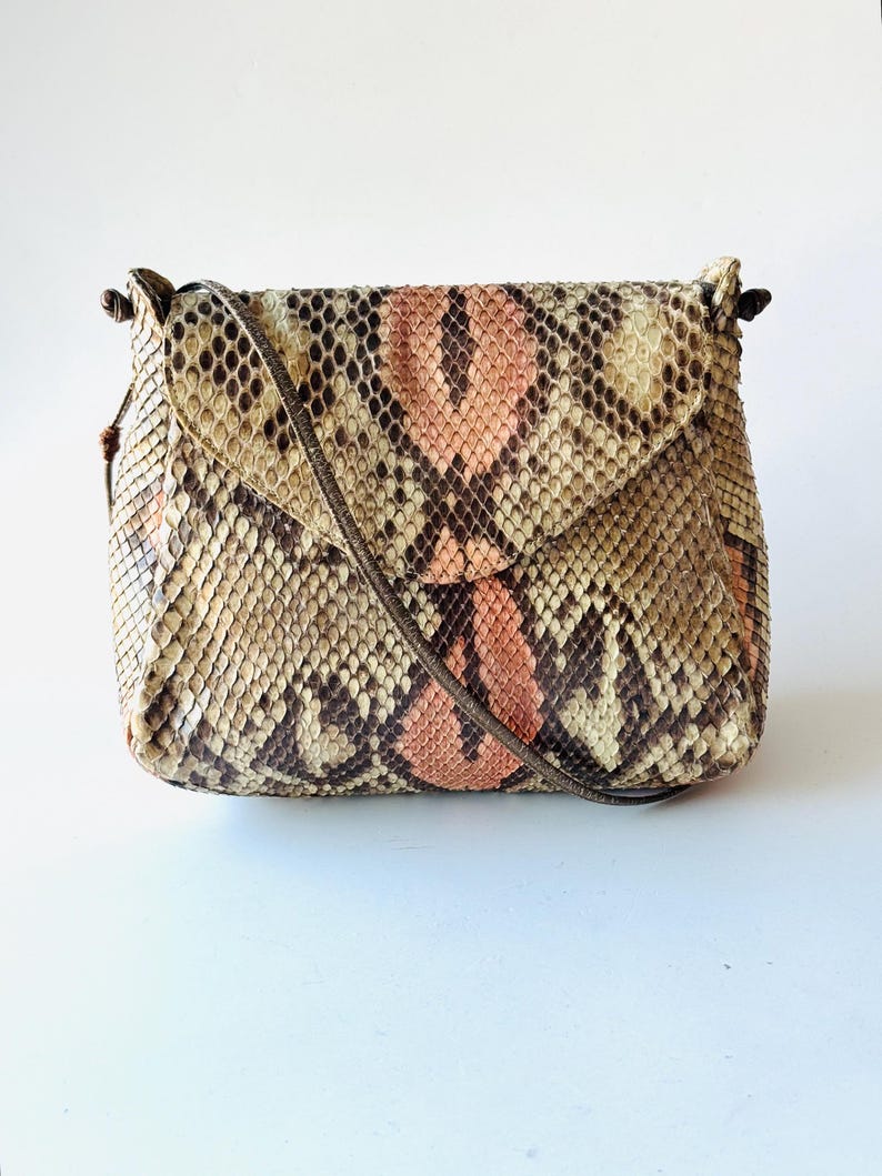 K&ouml;nnte beinhalten: Eine kleine, rechteckige Umh&auml;ngetasche mit Klappenverschluss. Die Tasche ist aus Schlangenhaut in den Farbt&ouml;nen Beige, Braun und Pfirsich gefertigt. Sie hat einen d&uuml;nnen, braunen Riemen und ein schlichtes, elegantes Design.