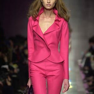 Pode incluir: Uma modelo com um fato rosa brilhante desfila numa passarela. O fato inclui um casaco justo com gola com folhos e cal&ccedil;as a condizer. A modelo carrega uma pequena clutch roxa escura. O fundo est&aacute; desfocado, com ilumina&ccedil;&atilde;o suave.