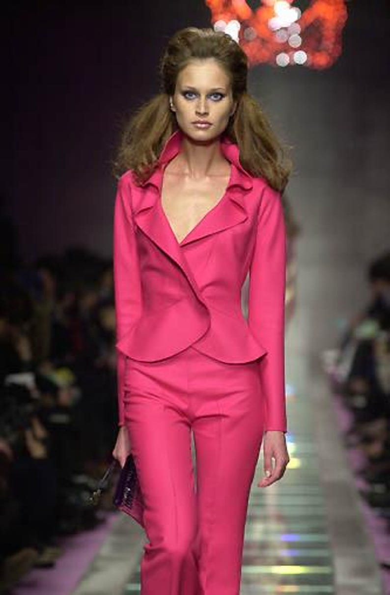 Pode incluir: Uma modelo com um fato rosa brilhante desfila numa passarela. O fato inclui um casaco justo com gola com folhos e cal&ccedil;as a condizer. A modelo carrega uma pequena clutch roxa escura. O fundo est&aacute; desfocado, com ilumina&ccedil;&atilde;o suave.