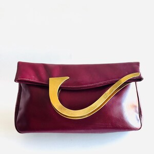 May include: A burgundy leather clutch with a gold-toned handle. The clutch has a rectangular shape with a fold-over flap. The handle is a curved, stylized design. The bag is set against a white background.