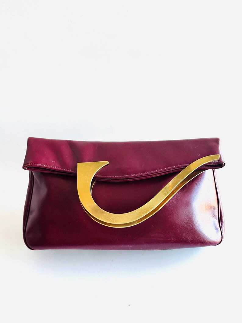 May include: A burgundy leather clutch with a gold-toned handle. The clutch has a rectangular shape with a fold-over flap. The handle is a curved, stylized design. The bag is set against a white background.