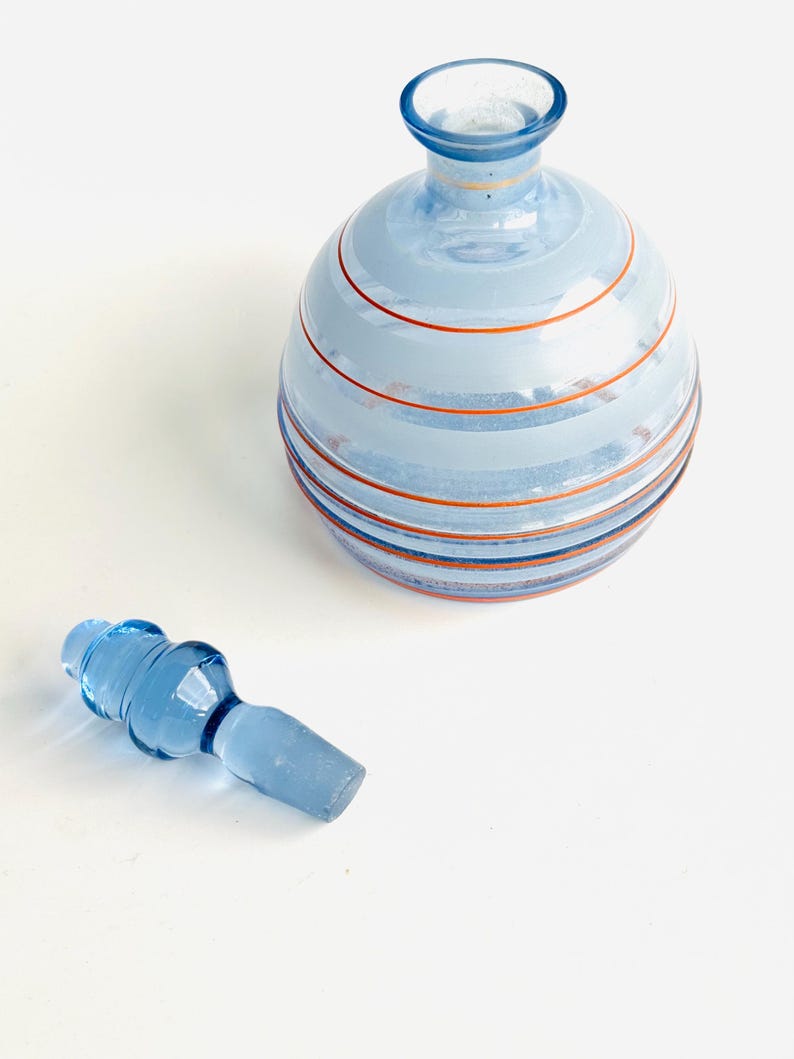 Pu&ograve; includere: Una bottiglia di vetro blu con un design a strisce e un tappo di vetro blu abbinato. La bottiglia ha un corpo rotondo e un collo stretto.