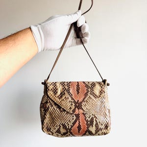 K&ouml;nnte beinhalten: Kleine, rechteckige Handtasche mit Klappenverschluss. Die Tasche ist aus Schlangenhaut in Braun-, Beige- und Rosat&ouml;nen gefertigt. Sie hat einen d&uuml;nnen, braunen Riemen und einen Schlaufengriff. Die Tasche wird von einer behandschuhten Hand gehalten.