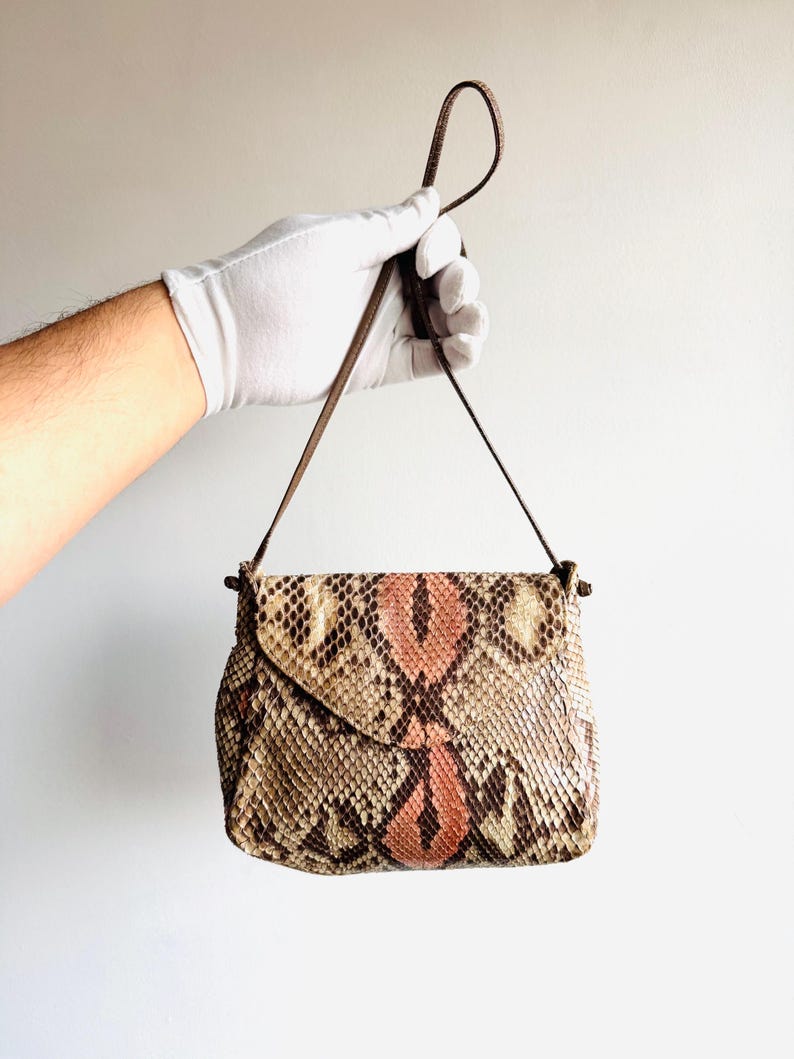 K&ouml;nnte beinhalten: Kleine, rechteckige Handtasche mit Klappenverschluss. Die Tasche ist aus Schlangenhaut in Braun-, Beige- und Rosat&ouml;nen gefertigt. Sie hat einen d&uuml;nnen, braunen Riemen und einen Schlaufengriff. Die Tasche wird von einer behandschuhten Hand gehalten.