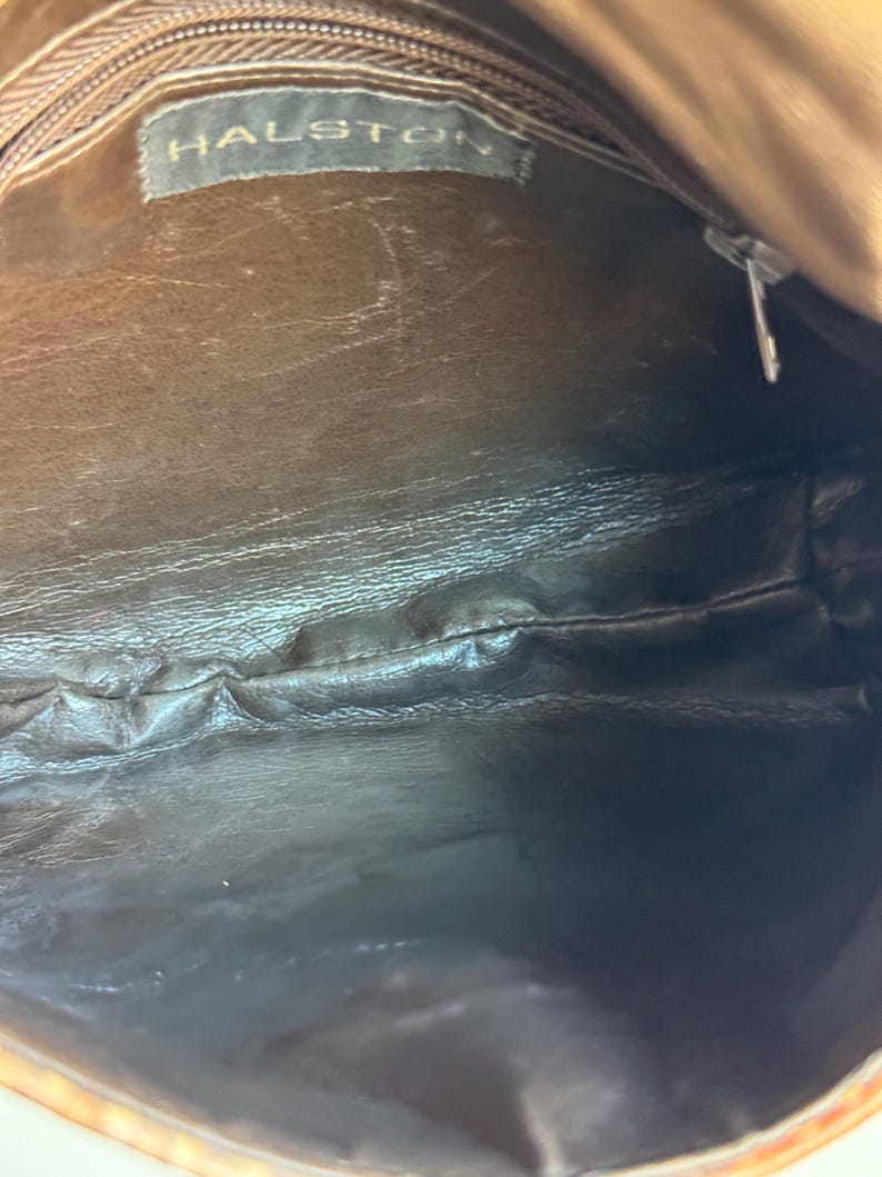 K&ouml;nnte beinhalten: Innenansicht einer braunen Lederhandtasche. Die Innenseite ist dunkelbraun mit schwarzem Boden. Der Markenname "HALSTON" ist auf einem Etikett im Inneren der Tasche zu sehen. Der Rei&szlig;verschluss ist silberfarben.