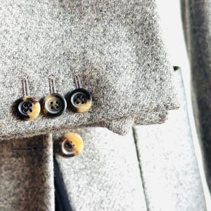 May include: Close-up of a gray wool coat with three buttons on the sleeve and one on the front. The buttons are brown and black with a marbled effect. The coat has a classic design.