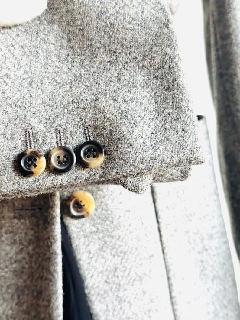 May include: Close-up of a gray wool coat with three buttons on the sleeve and one on the front. The buttons are brown and black with a marbled effect. The coat has a classic design.