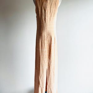 May include: A peach-colored, sleeveless maxi dress with a subtle textured pattern. The dress features a scoop neckline and a back zipper. A slit runs up the back of the dress. The dress is displayed on a mannequin.