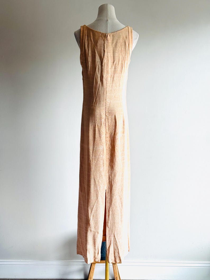 May include: A peach-colored, sleeveless maxi dress with a subtle textured pattern. The dress features a scoop neckline and a back zipper. A slit runs up the back of the dress. The dress is displayed on a mannequin.