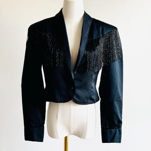 May include: Black satin jacket with beaded fringe detail across the shoulders. The jacket has a notched collar, a single button closure, and long sleeves with buttoned cuffs. The hemline has a scalloped edge.