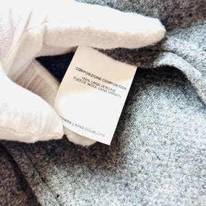 May include: Close-up of a gray fabric with a white tag. The tag displays text in multiple languages, including "COMPOSIZIONE-COMPOSITION" and "100% LANA VERGINE FLEECE WOOL LANA VIRGEN". The fabric has a textured appearance.