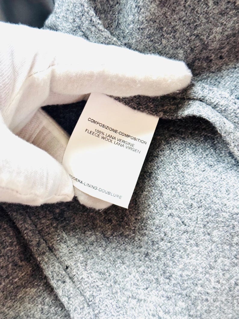 May include: Close-up of a gray fabric with a white tag. The tag displays text in multiple languages, including "COMPOSIZIONE-COMPOSITION" and "100% LANA VERGINE FLEECE WOOL LANA VIRGEN". The fabric has a textured appearance.