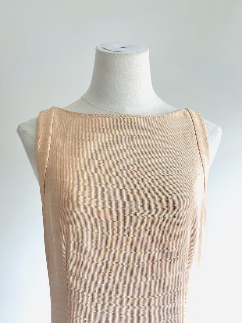 May include: A sleeveless, peach-colored dress with a textured, ribbed fabric. The dress has a boat neckline and is displayed on a mannequin. The fabric has a subtle, horizontal wave pattern.