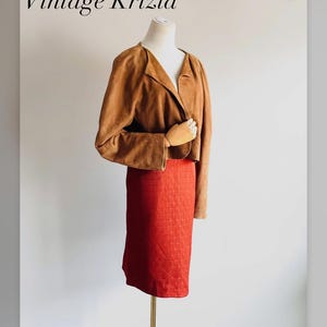 May include: A tan suede jacket with long sleeves and a V-neckline paired with a textured red knee-length skirt. The image includes the text Vintage Krizia at the top and @foreveragainshop at the bottom. The garments are displayed on a mannequin.