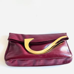 May include: A burgundy leather handbag with a large, curved gold-tone handle. The bag has a fold-over flap and a rectangular shape. The leather appears slightly worn, adding a vintage aesthetic. The handle is a prominent design element.