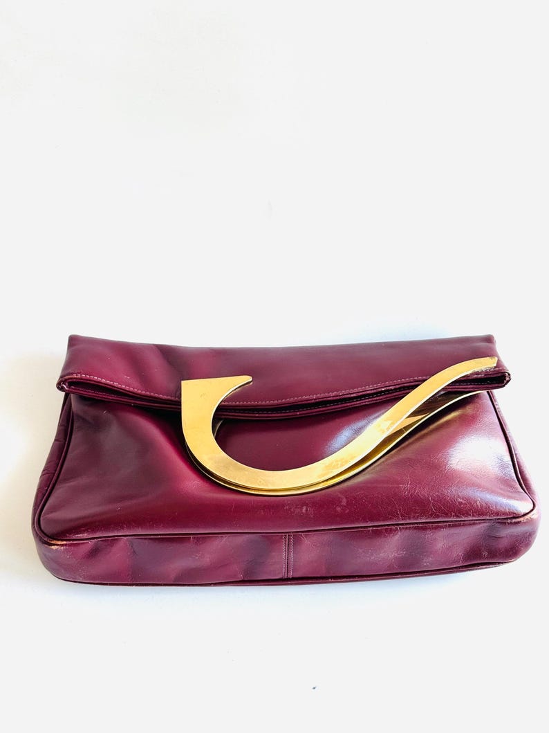 May include: A burgundy leather handbag with a large, curved gold-tone handle. The bag has a fold-over flap and a rectangular shape. The leather appears slightly worn, adding a vintage aesthetic. The handle is a prominent design element.