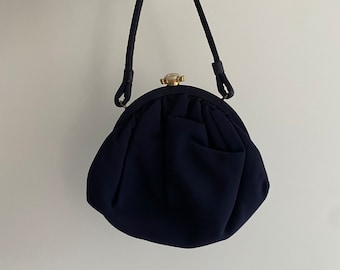 May include: A small, dark blue, round purse with a gold clasp and a short handle. The purse is made of a soft, textured fabric.