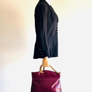 May include: A black blazer with silver buttons and a burgundy leather handbag with gold-tone handles. The blazer is displayed on a mannequin. The handbag is a structured tote style.