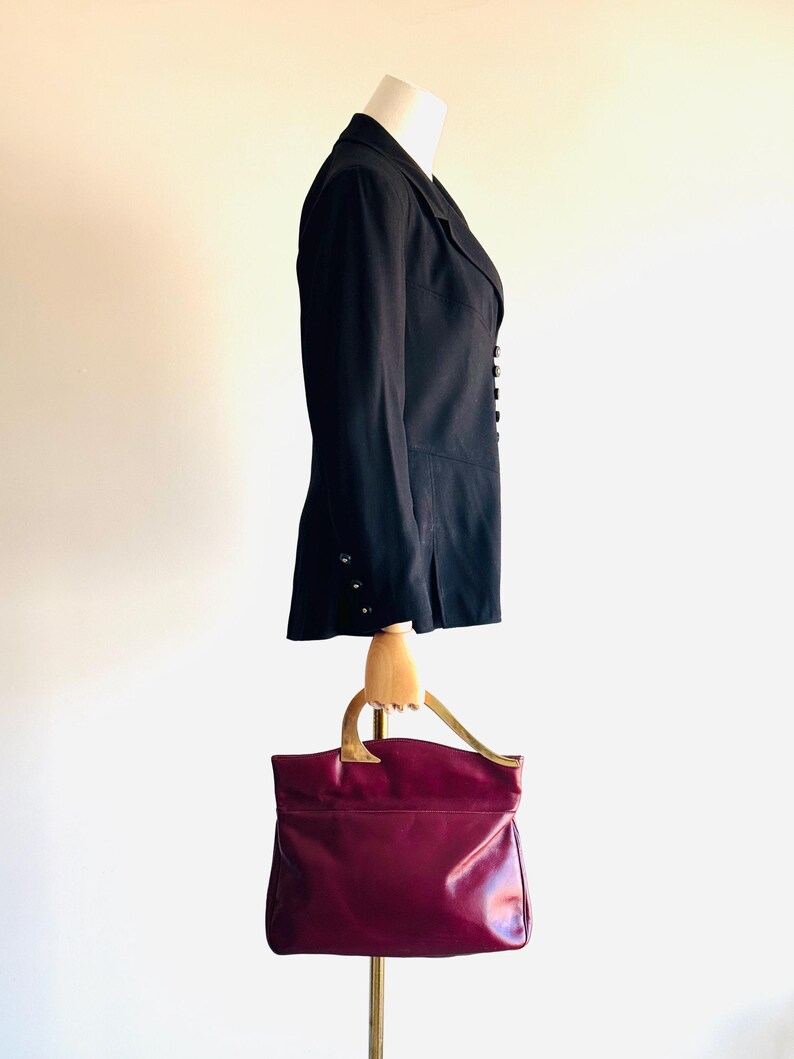 May include: A black blazer with silver buttons and a burgundy leather handbag with gold-tone handles. The blazer is displayed on a mannequin. The handbag is a structured tote style.