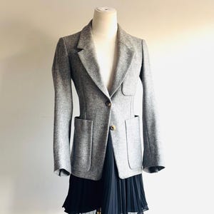 May include: A gray blazer with a tailored fit, featuring two front pockets and a single-button closure. The blazer is displayed on a mannequin, paired with a black pleated skirt. The outfit is set against a neutral background.