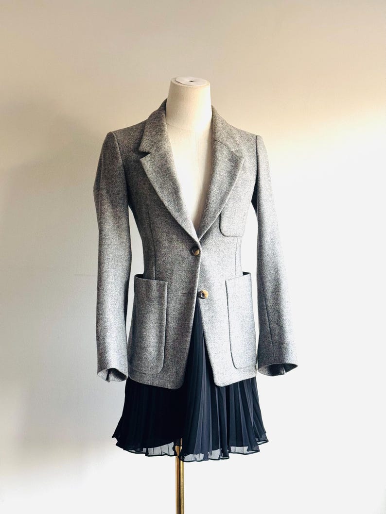 May include: A gray blazer with a tailored fit, featuring two front pockets and a single-button closure. The blazer is displayed on a mannequin, paired with a black pleated skirt. The outfit is set against a neutral background.