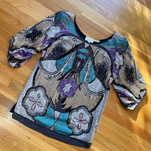 May include: A black sequined top with a colorful floral pattern. The top has a scoop neck and short sleeves. The sequins are in shades of gold, silver, blue, purple, and pink.