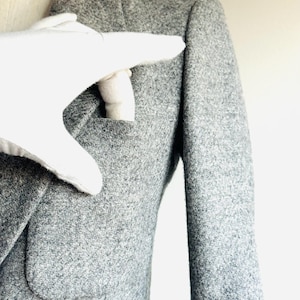 May include: A close-up of a gray blazer with a textured fabric. The blazer has a notched lapel, a pocket, and a sleeve. A white glove is touching the blazer's pocket.