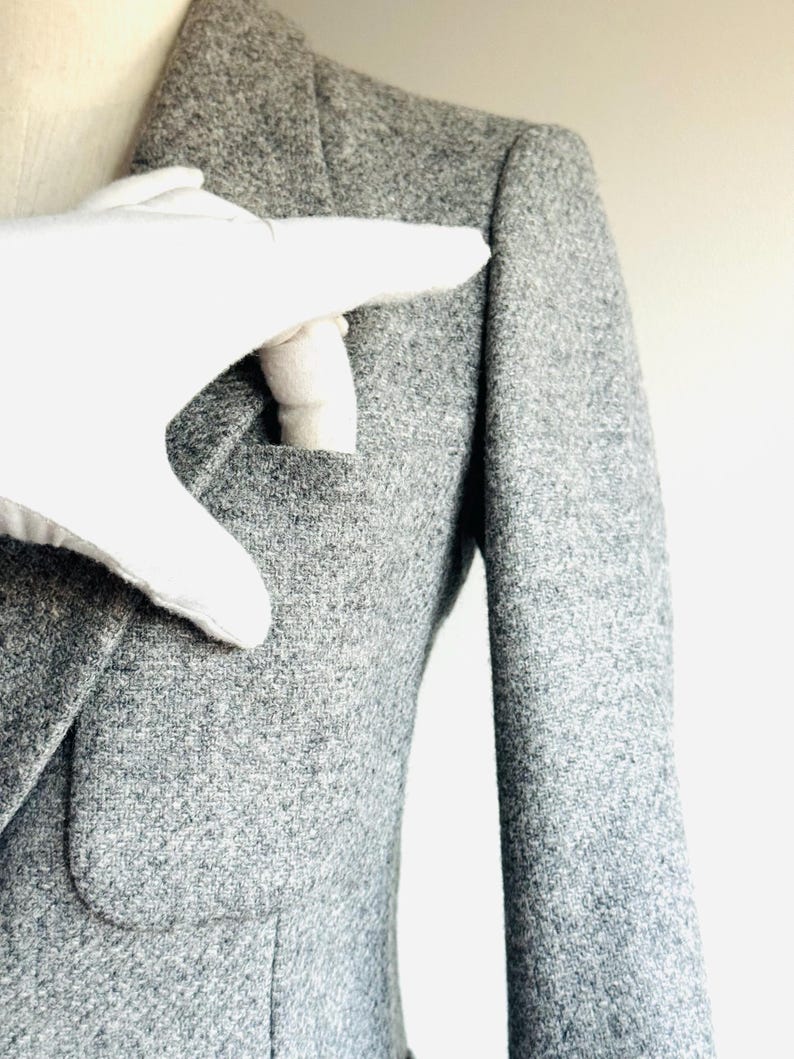 May include: A close-up of a gray blazer with a textured fabric. The blazer has a notched lapel, a pocket, and a sleeve. A white glove is touching the blazer's pocket.
