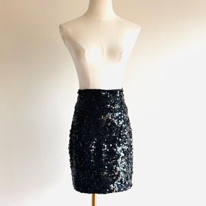 May include: A black sequin mini skirt displayed on a mannequin. The skirt is covered in small, shiny sequins that reflect light. The mannequin is a neutral color, and the skirt is the focal point.