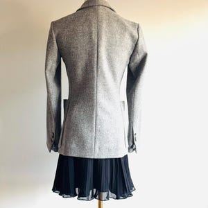 May include: A light gray blazer with a classic design, displayed on a mannequin. The blazer has a tailored fit, long sleeves, and a notched lapel. A black pleated skirt is worn below the blazer. The outfit is displayed on a gold-colored stand.