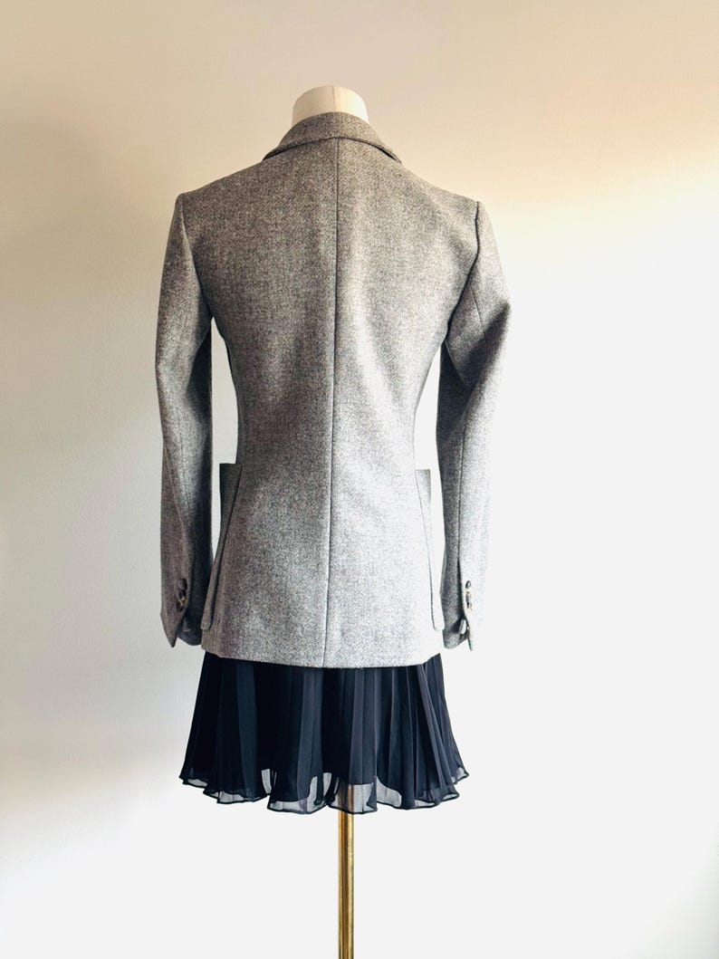 May include: A light gray blazer with a classic design, displayed on a mannequin. The blazer has a tailored fit, long sleeves, and a notched lapel. A black pleated skirt is worn below the blazer. The outfit is displayed on a gold-colored stand.