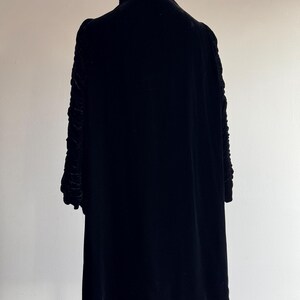 K&ouml;nnte beinhalten: Ein schwarzes Samtkleid mit hohem Kragen und langen &Auml;rmeln. Die &Auml;rmel haben ein gerafftes Detail.
