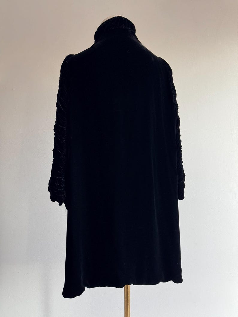 K&ouml;nnte beinhalten: Ein schwarzes Samtkleid mit hohem Kragen und langen &Auml;rmeln. Die &Auml;rmel haben ein gerafftes Detail.
