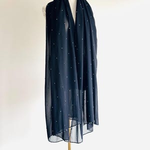 May include: A long, rectangular black scarf made of a sheer, lightweight fabric. The scarf is decorated with evenly spaced, small white pearl-like embellishments scattered across the entire surface, creating a subtle, textured, and dotted appearance.