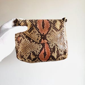 K&ouml;nnte beinhalten: Eine kleine, rechteckige Clutch mit einem Schlangenmuster in Braun-, Beige- und Pfirsicht&ouml;nen. Die Clutch hat einen Klappenverschluss und ein subtiles, strukturiertes Aussehen. Das Gesamtdesign ist kompakt und stilvoll, geeignet f&uuml;r das Tragen von Essentials.